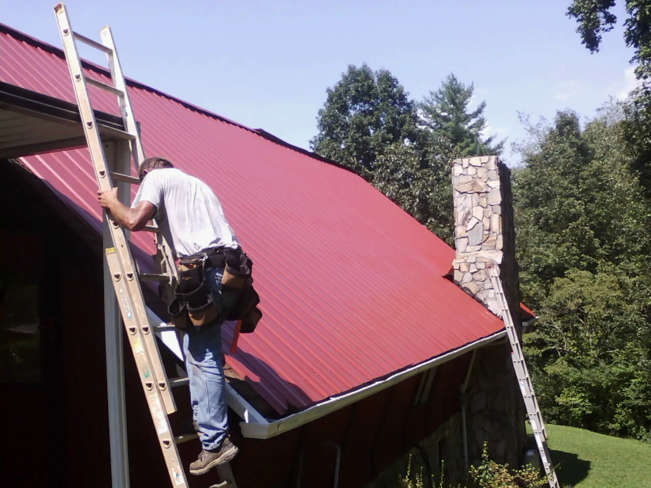 Roofer on a ladder beside a red metal roof and stone chimney — Soffit & Fascia Repair by Roofing Brothers in Eustis